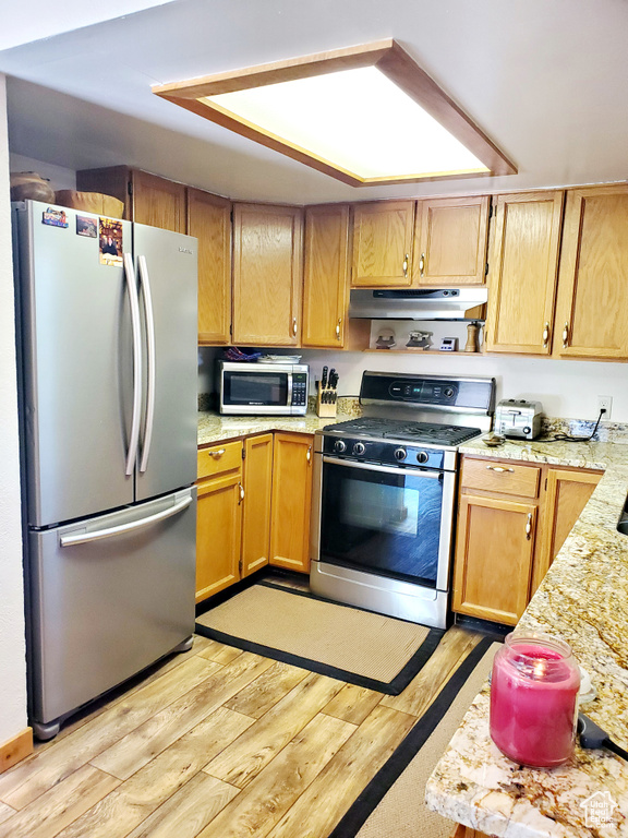 Kitchen with stainless steel appliances, under cabinet range hood, light wood-style flooring, and light stone counters