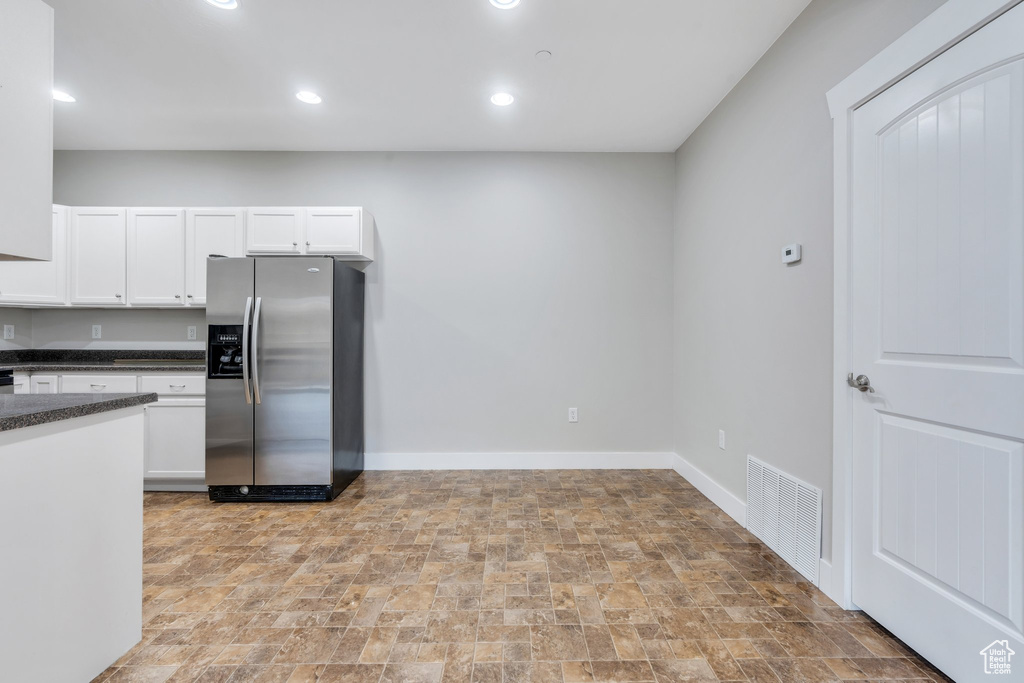 Kitchen with stainless steel refrigerator with ice dispenser, dark countertops, stone finish floors, white cabinetry, and baseboards