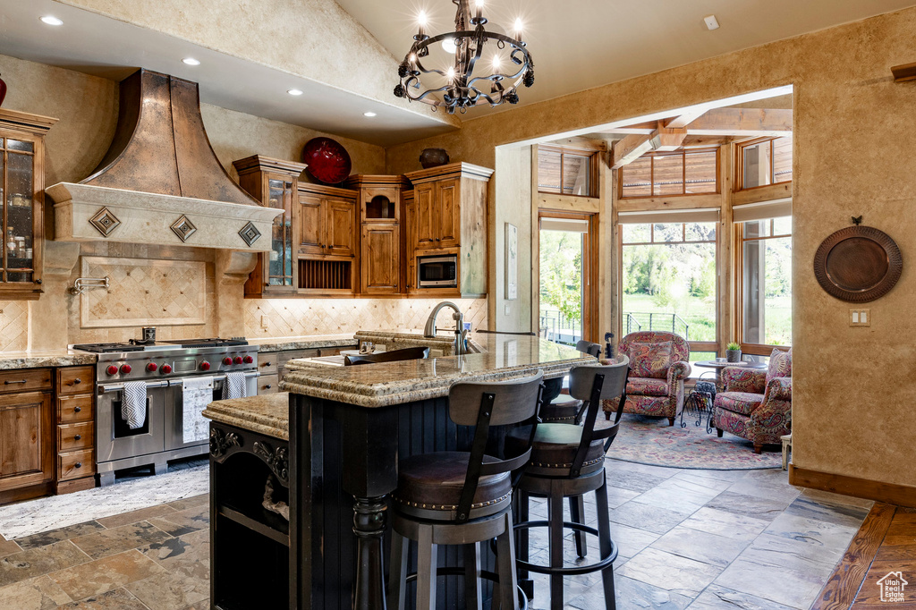 Kitchen featuring stainless steel appliances, premium range hood, brown cabinetry, decorative backsplash, and stone tile floors