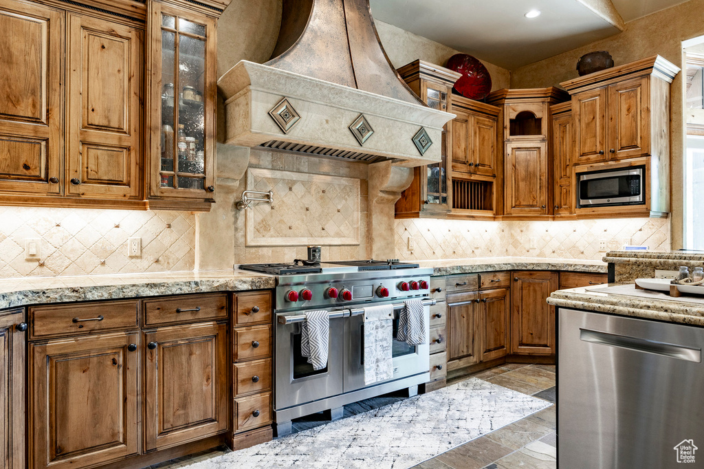 Kitchen with appliances with stainless steel finishes, custom range hood, brown cabinets, backsplash, and recessed lighting
