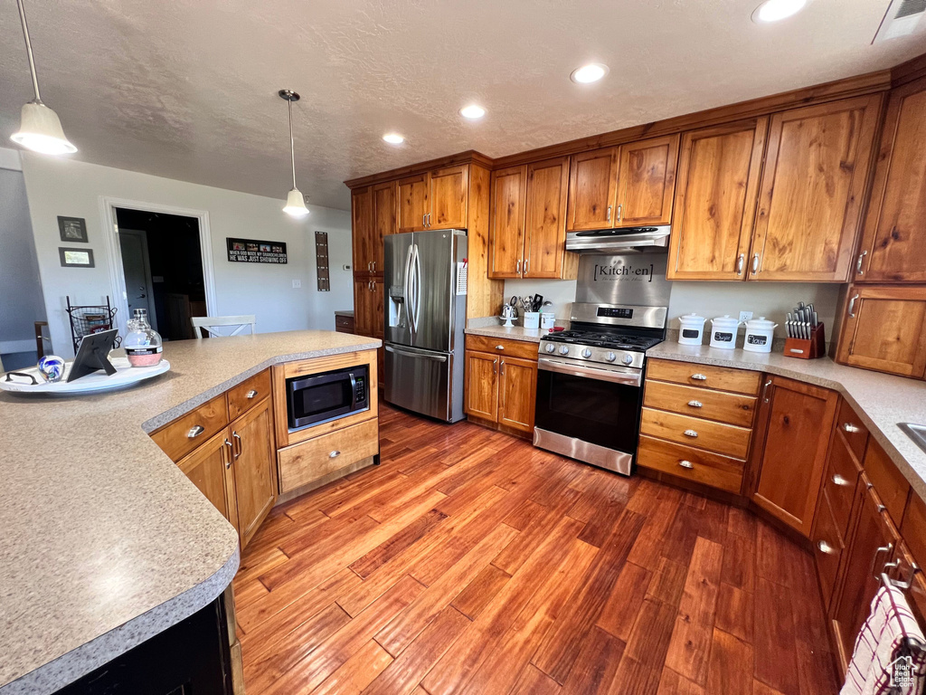 Kitchen with stainless steel appliances, under cabinet range hood, wood finished floors, brown cabinetry, and recessed lighting