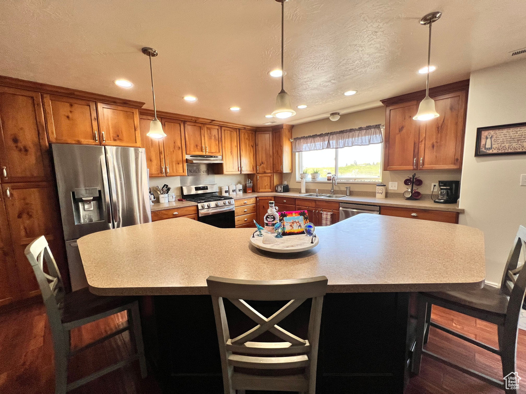 Kitchen featuring appliances with stainless steel finishes, under cabinet range hood, a breakfast bar, brown cabinetry, and recessed lighting