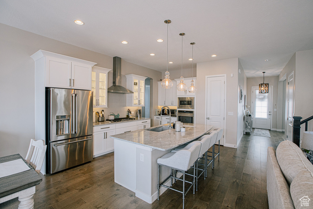 Kitchen featuring appliances with stainless steel finishes, wall chimney exhaust hood, a sink, a kitchen island with sink, and a breakfast bar area