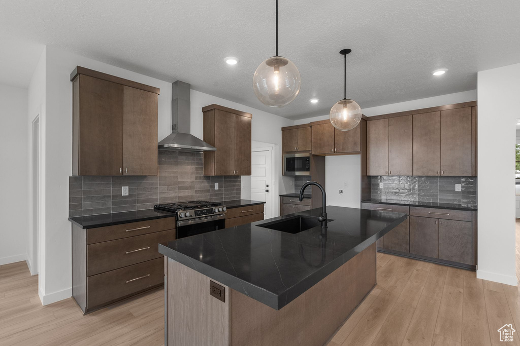 Kitchen featuring wall chimney range hood, a sink, dark countertops, appliances with stainless steel finishes, and light wood-style floors