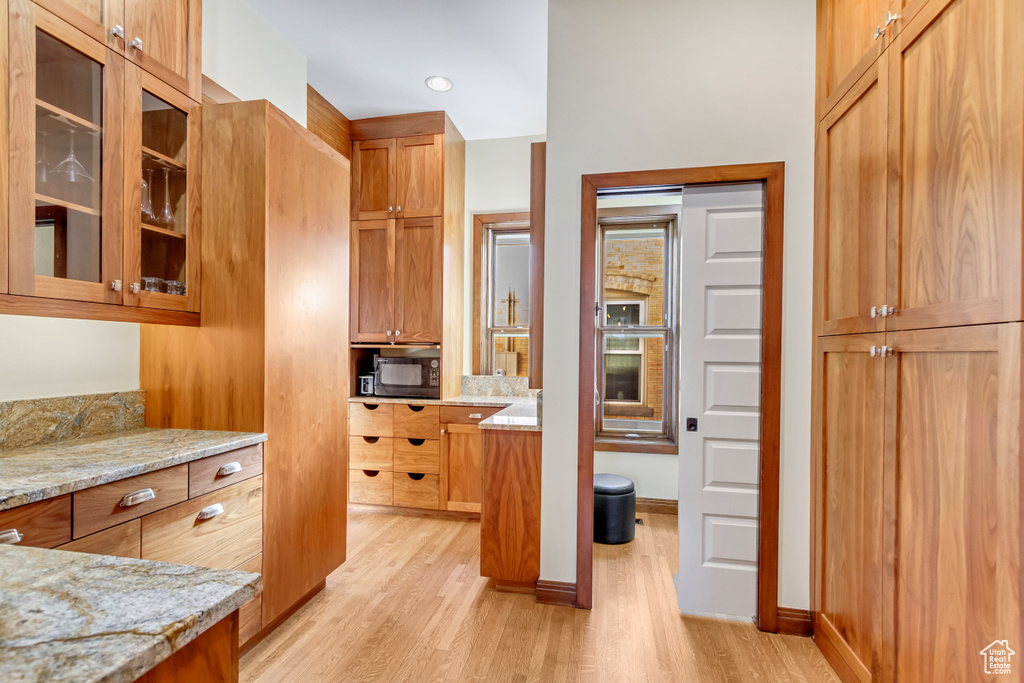 Kitchen with light wood-style flooring, light stone counters, glass insert cabinets, and brown cabinetry