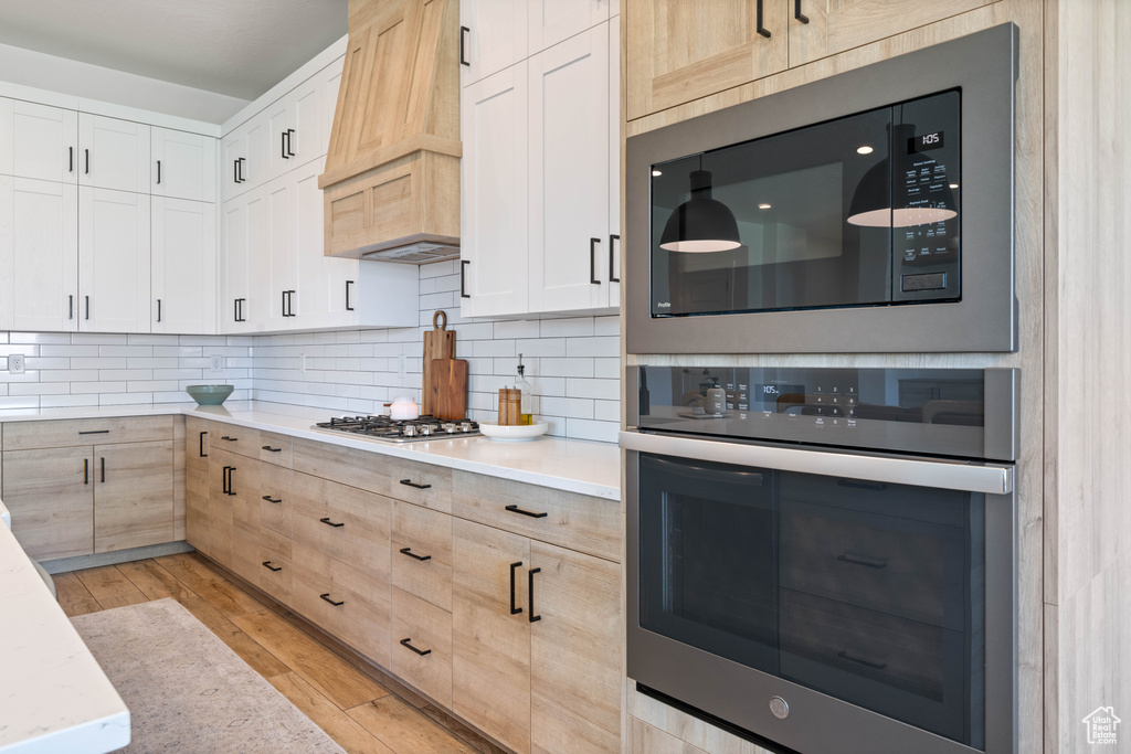 Kitchen with appliances with stainless steel finishes, custom exhaust hood, decorative backsplash, light brown cabinets, and light wood finished floors
