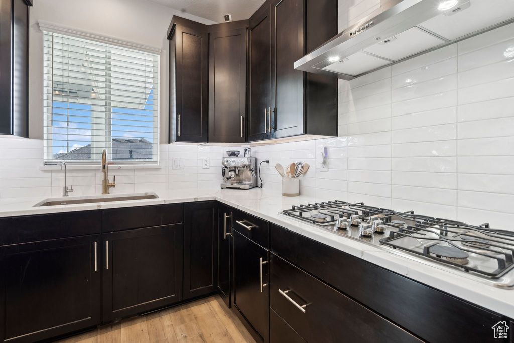 Kitchen featuring wall chimney exhaust hood, a sink, stainless steel gas cooktop, decorative backsplash, and light wood-style flooring