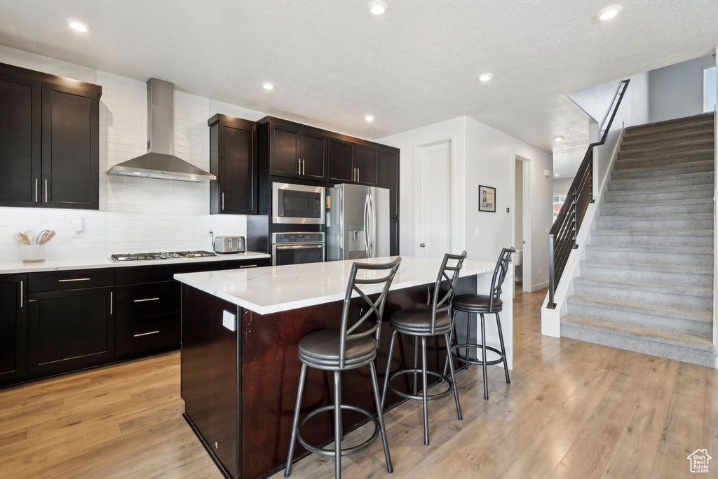 Kitchen with wall chimney range hood, stainless steel appliances, a kitchen breakfast bar, a center island, and light wood finished floors