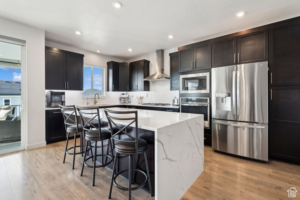 Kitchen featuring appliances with stainless steel finishes, wall chimney exhaust hood, light stone countertops, a center island, and a breakfast bar