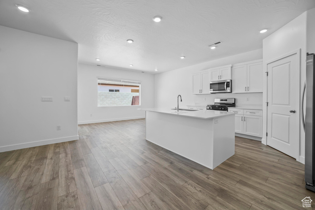 Kitchen featuring stainless steel appliances, a sink, white cabinetry, wood finished floors, and a kitchen island with sink