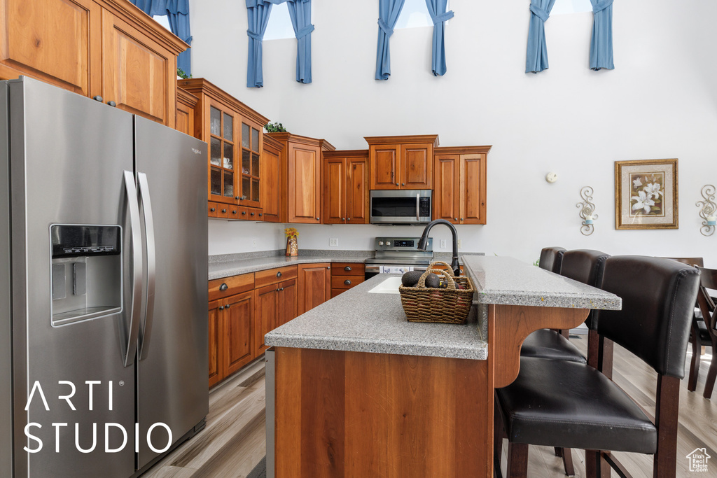 Kitchen featuring stainless steel appliances, a towering ceiling, brown cabinets, light countertops, and an island with sink
