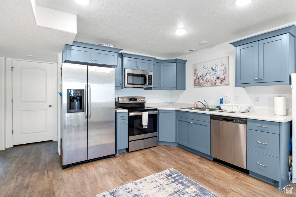Kitchen with appliances with stainless steel finishes, a sink, light countertops, light wood-type flooring, and a textured ceiling