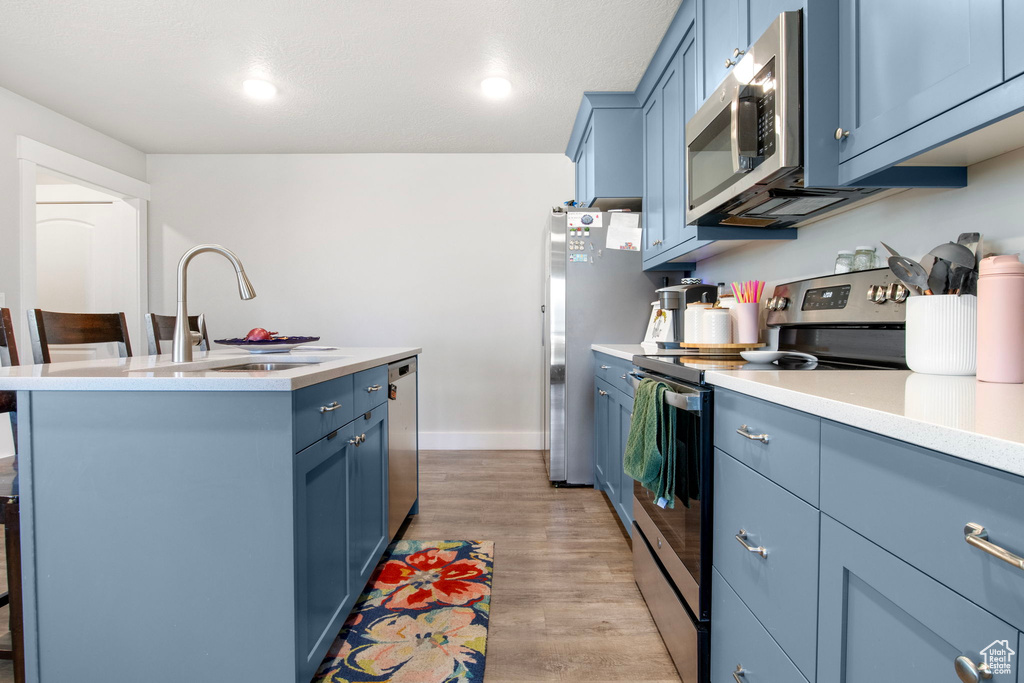 Kitchen with stainless steel appliances, blue cabinetry, a sink, light countertops, and an island with sink