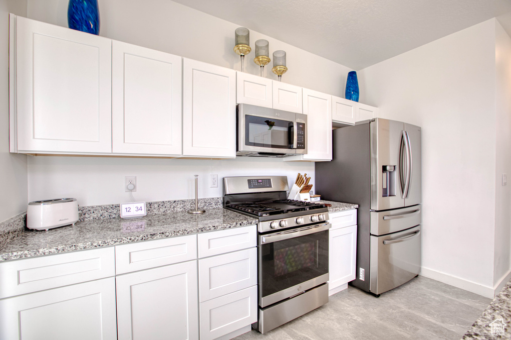 Kitchen with appliances with stainless steel finishes, white cabinetry, light stone counters, light wood-type flooring, and baseboards
