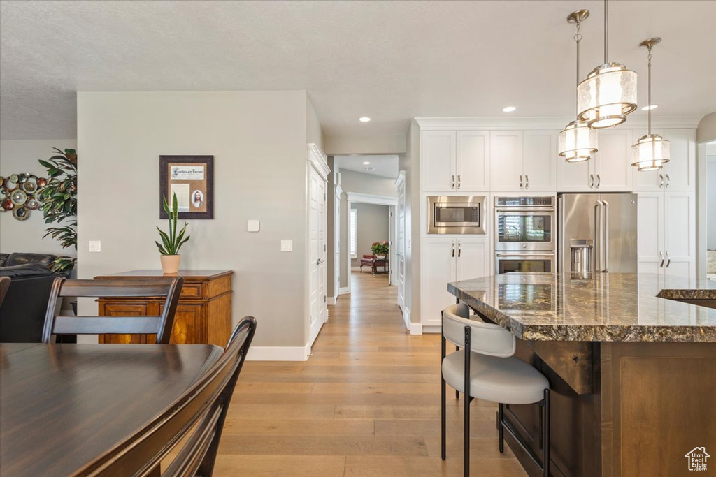 Kitchen featuring appliances with stainless steel finishes, light wood-style flooring, dark stone countertops, white cabinetry, and hanging light fixtures