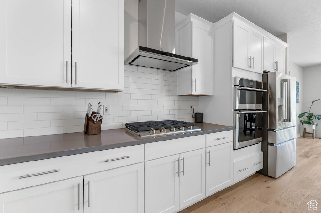 Kitchen with wall chimney range hood, stainless steel appliances, white cabinetry, dark countertops, and light wood-style floors