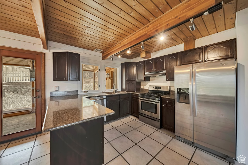 Kitchen with appliances with stainless steel finishes, a sink, wooden ceiling with exposed beams, dark stone countertops, and light tile patterned flooring