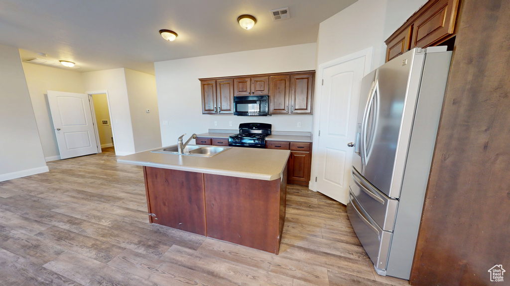 Kitchen featuring black appliances, a sink, light wood-type flooring, a kitchen island with sink, and light countertops