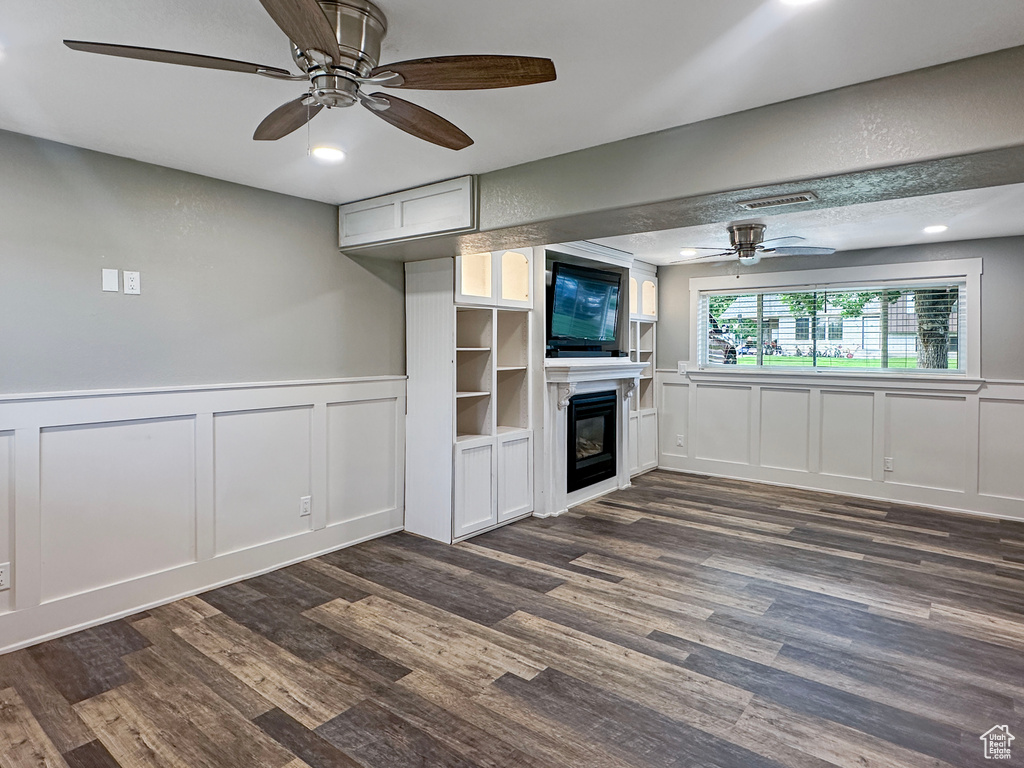 Unfurnished living room featuring a decorative wall, a ceiling fan, a glass covered fireplace, wainscoting, and dark wood finished floors