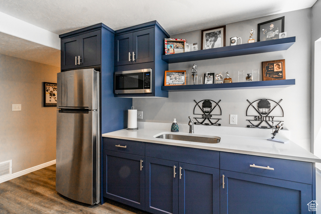 Kitchen with appliances with stainless steel finishes, a sink, open shelves, blue cabinetry, and dark wood-style floors