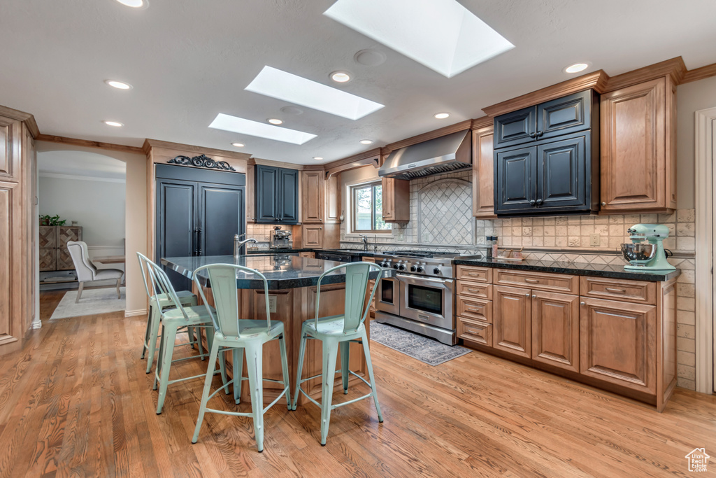Kitchen featuring range with two ovens, wall chimney exhaust hood, a skylight, a breakfast bar area, and light wood-style floors