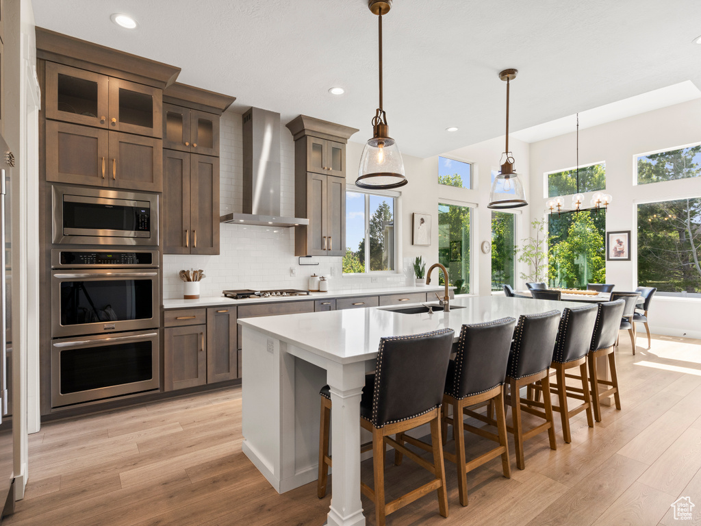 Kitchen with a sink, wall chimney range hood, appliances with stainless steel finishes, light countertops, and light wood-style flooring