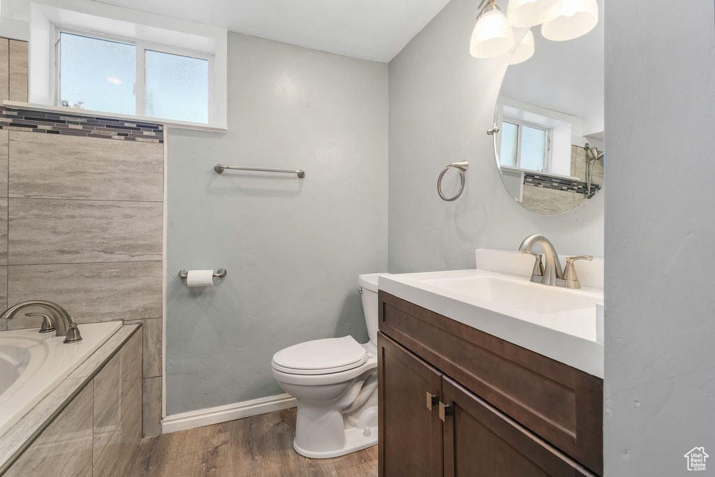 Bathroom featuring wood finished floors, toilet, a relaxing tiled tub, vanity, and baseboards