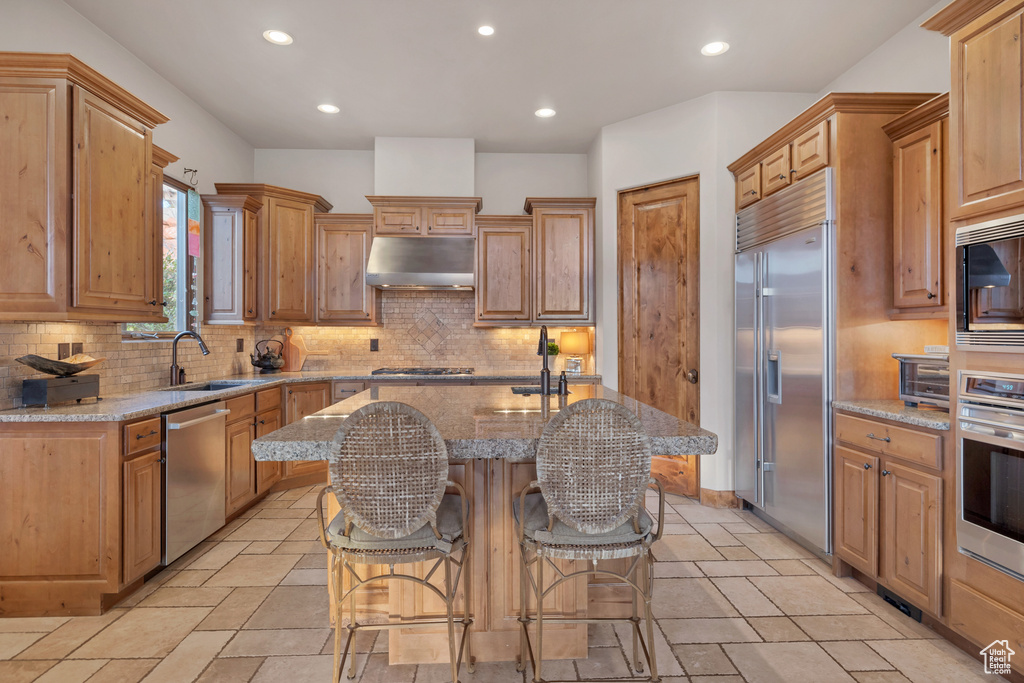 Kitchen with an island with sink, light stone counters, built in appliances, under cabinet range hood, and a kitchen bar