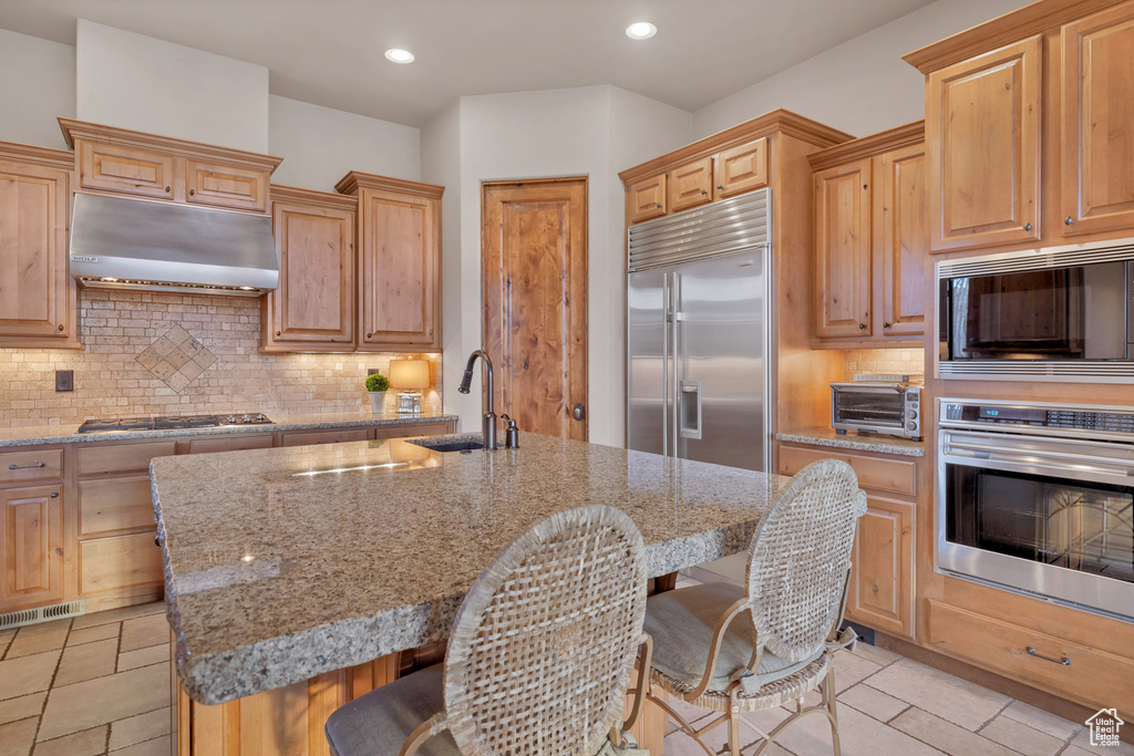 Kitchen with light stone counters, a breakfast bar, a sink, built in appliances, and under cabinet range hood