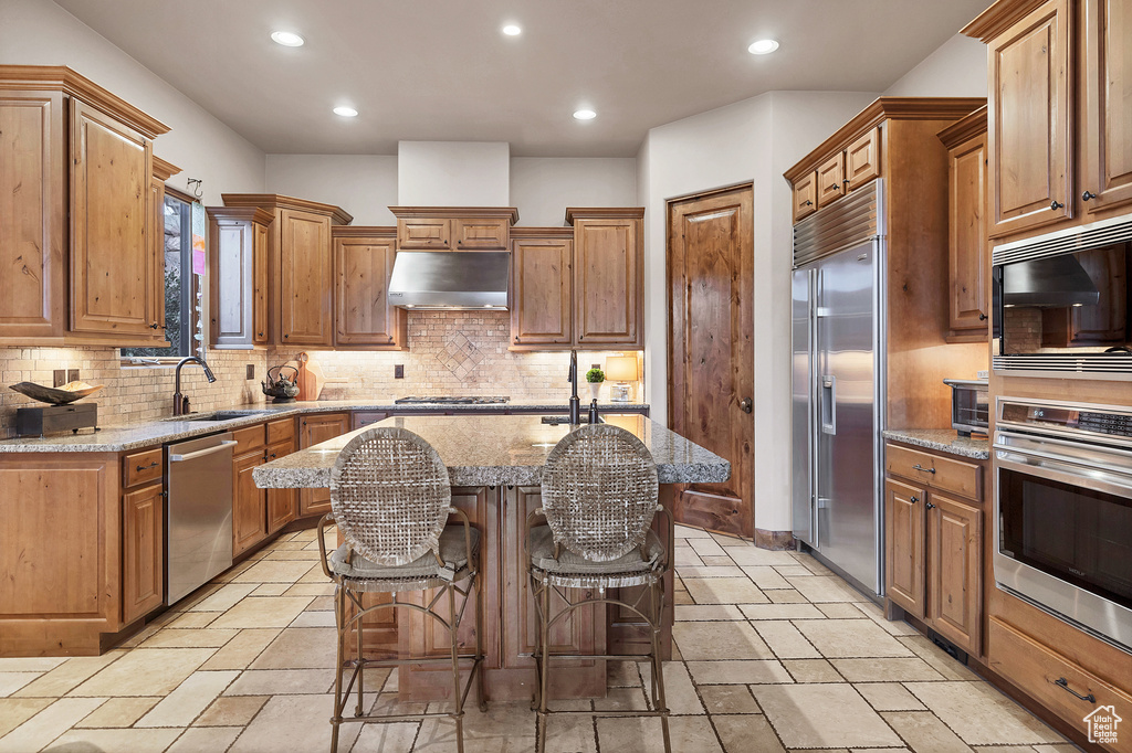 Kitchen featuring a center island with sink, a sink, light stone countertops, under cabinet range hood, and built in appliances
