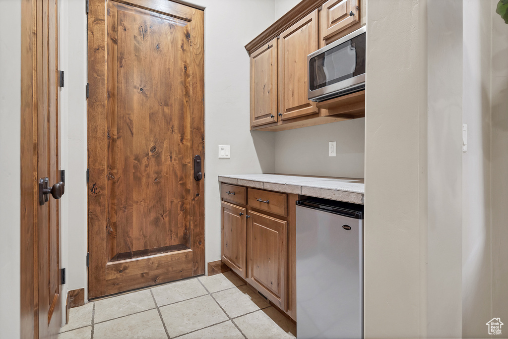 Kitchen with light tile patterned floors, tile countertops, stainless steel microwave, and fridge