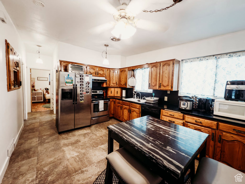 Kitchen featuring stainless steel appliances, under cabinet range hood, brown cabinets, dark countertops, and ceiling fan
