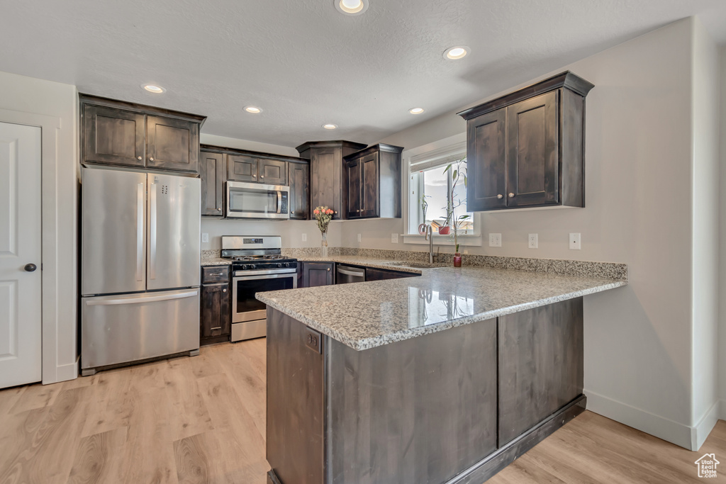 Kitchen with stainless steel appliances, a peninsula, dark brown cabinets, light wood finished floors, and light stone countertops