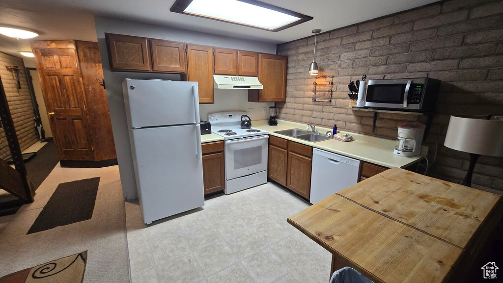 Kitchen with white appliances, a sink, brick wall, under cabinet range hood, and hanging light fixtures
