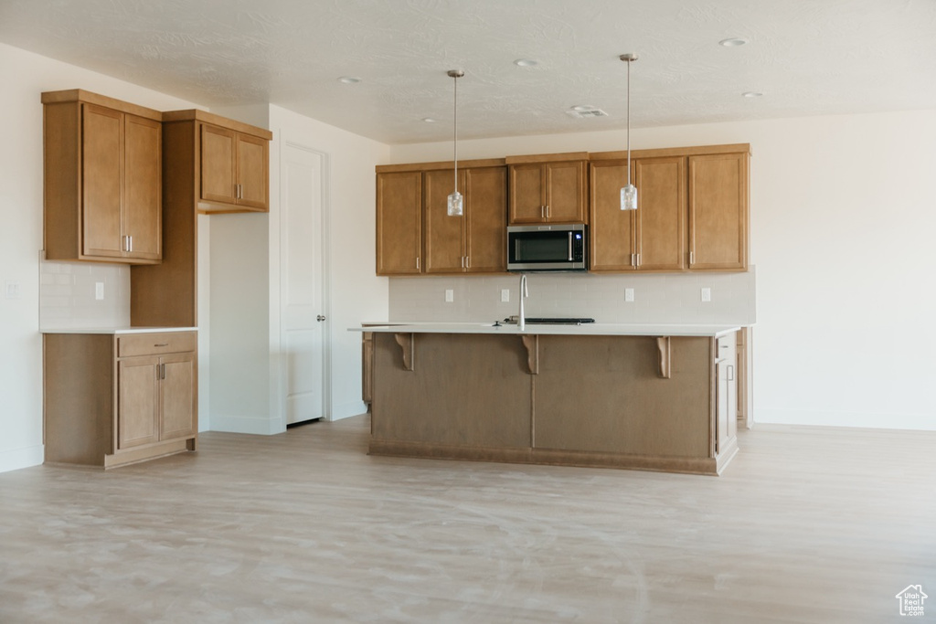 Kitchen with stainless steel microwave, tasteful backsplash, brown cabinets, a kitchen island with sink, and light countertops