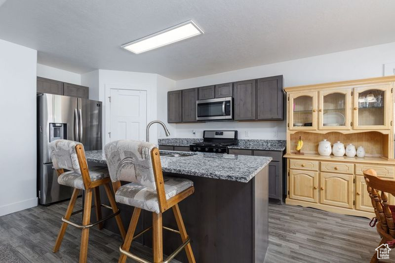 Kitchen with appliances with stainless steel finishes, a kitchen bar, glass insert cabinets, a kitchen island with sink, and dark wood-style floors