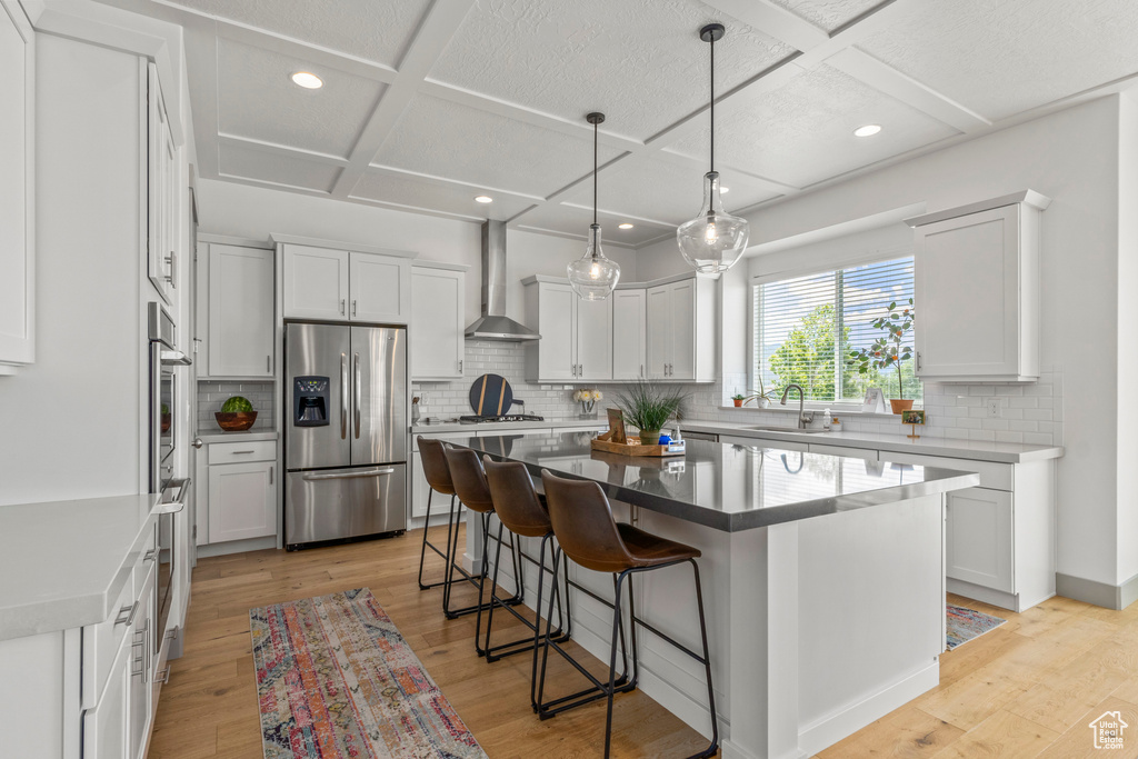 Kitchen featuring stainless steel fridge with ice dispenser, coffered ceiling, wall chimney range hood, a sink, and a center island