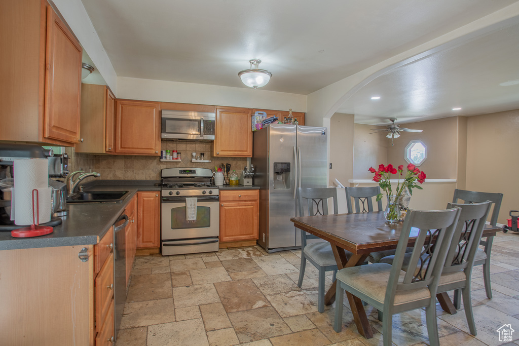 Kitchen with stainless steel appliances, a sink, ceiling fan, dark countertops, and stone tile flooring