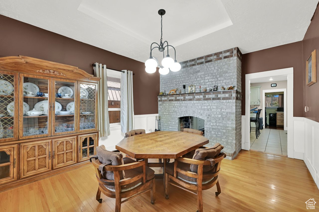 Dining space featuring a brick fireplace, light wood finished floors, wainscoting, a chandelier, and a tray ceiling