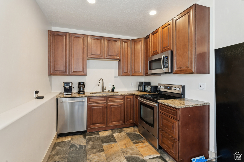 Kitchen featuring appliances with stainless steel finishes, a sink, stone finish flooring, light stone countertops, and brown cabinetry