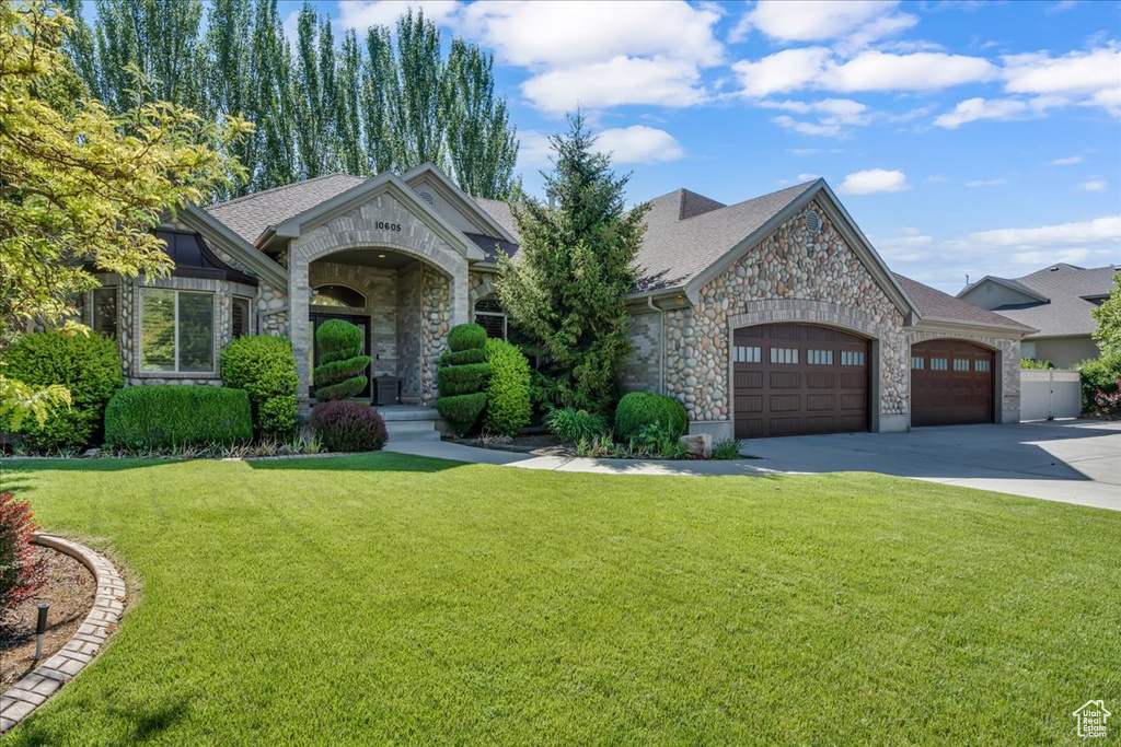 View of front of property with an attached garage, stone siding, concrete driveway, a front yard, and roof with shingles