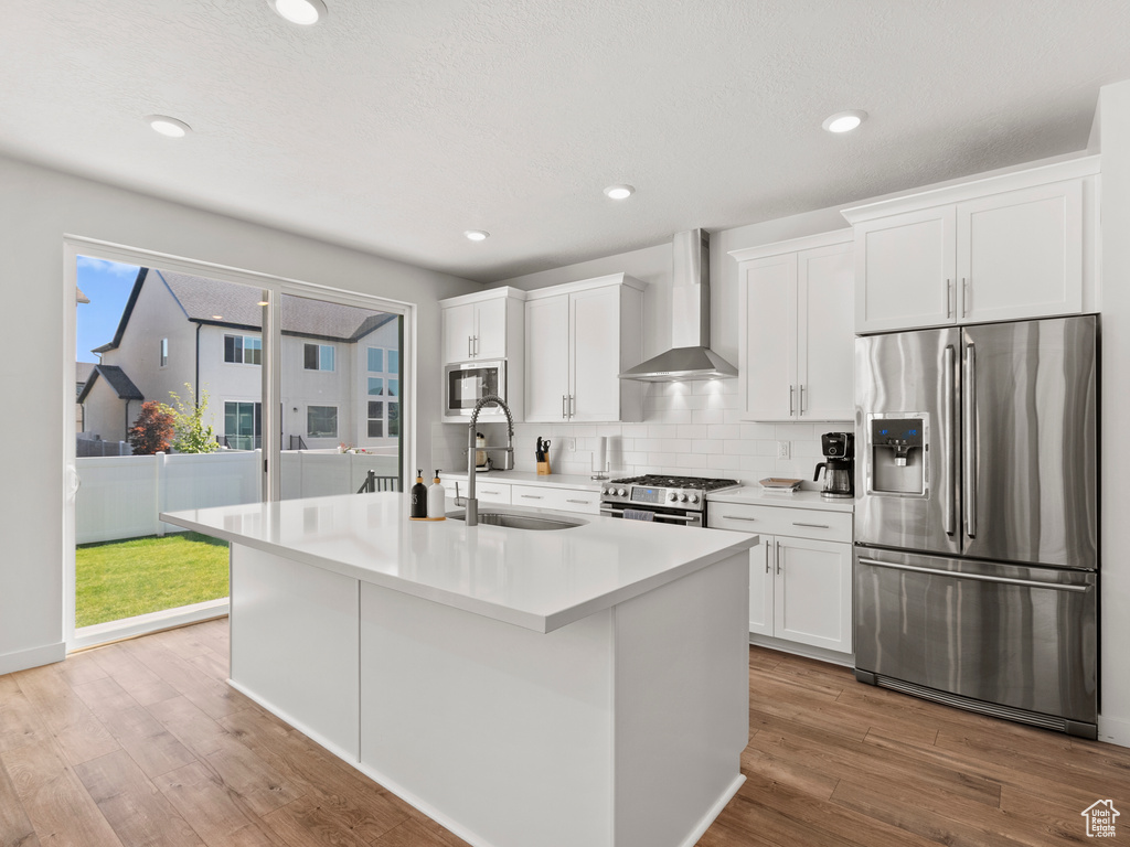 Kitchen featuring appliances with stainless steel finishes, wall chimney range hood, a sink, white cabinets, and an island with sink
