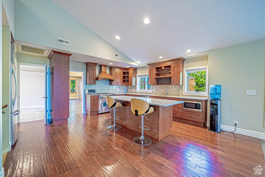Kitchen with open shelves, appliances with stainless steel finishes, custom exhaust hood, brown cabinetry, and dark wood-style flooring