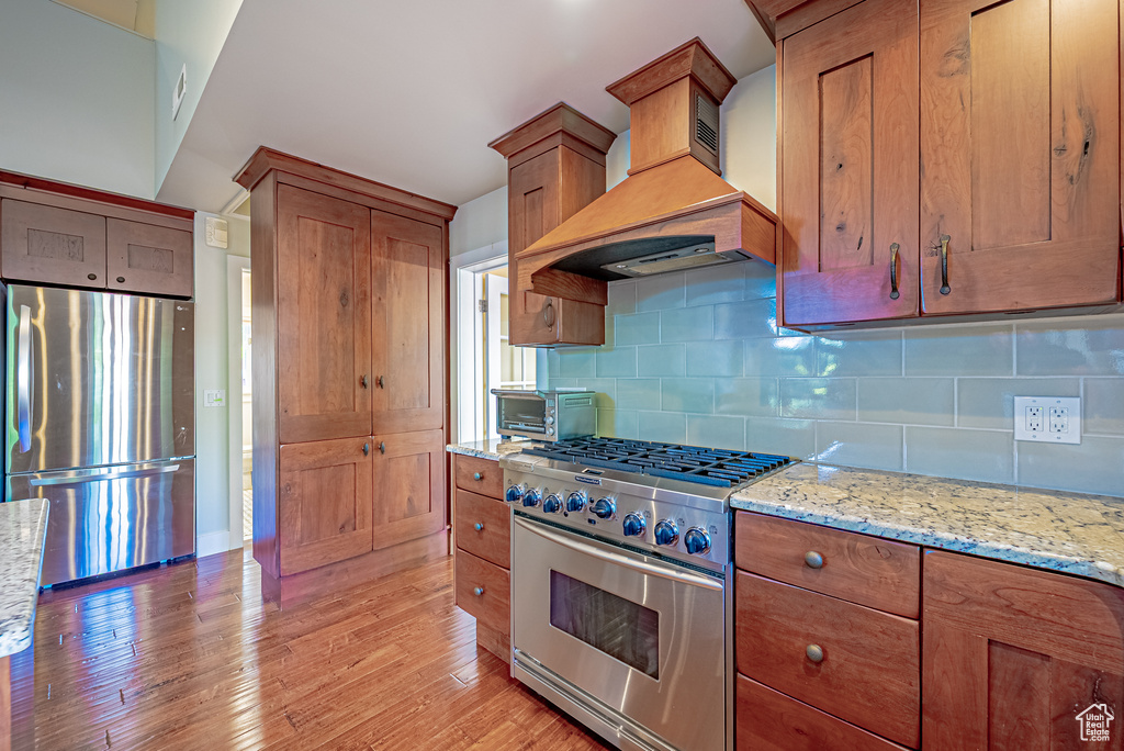 Kitchen with appliances with stainless steel finishes, custom range hood, brown cabinetry, and decorative backsplash