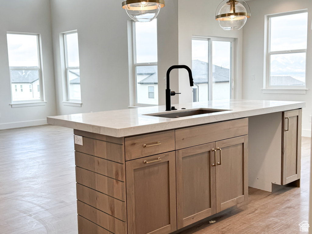 Kitchen with a sink, a center island with sink, light wood-style flooring, light stone counters, and decorative light fixtures