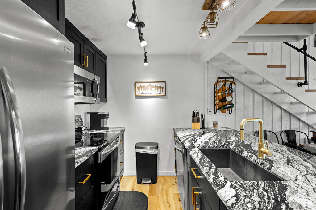 Kitchen featuring stainless steel appliances, a sink, dark cabinetry, light wood-style flooring, and light stone countertops