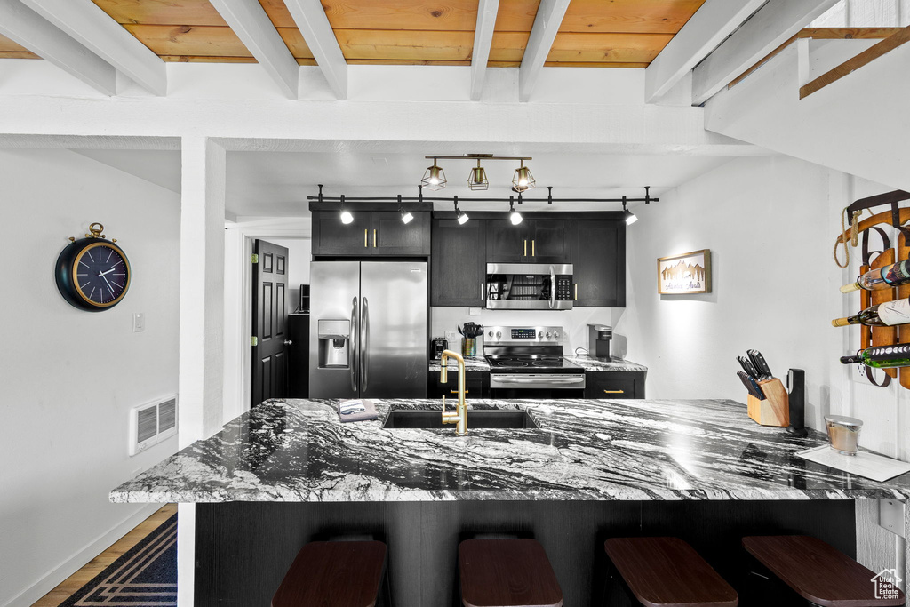 Kitchen with stainless steel appliances, dark cabinetry, a kitchen breakfast bar, a sink, and beam ceiling