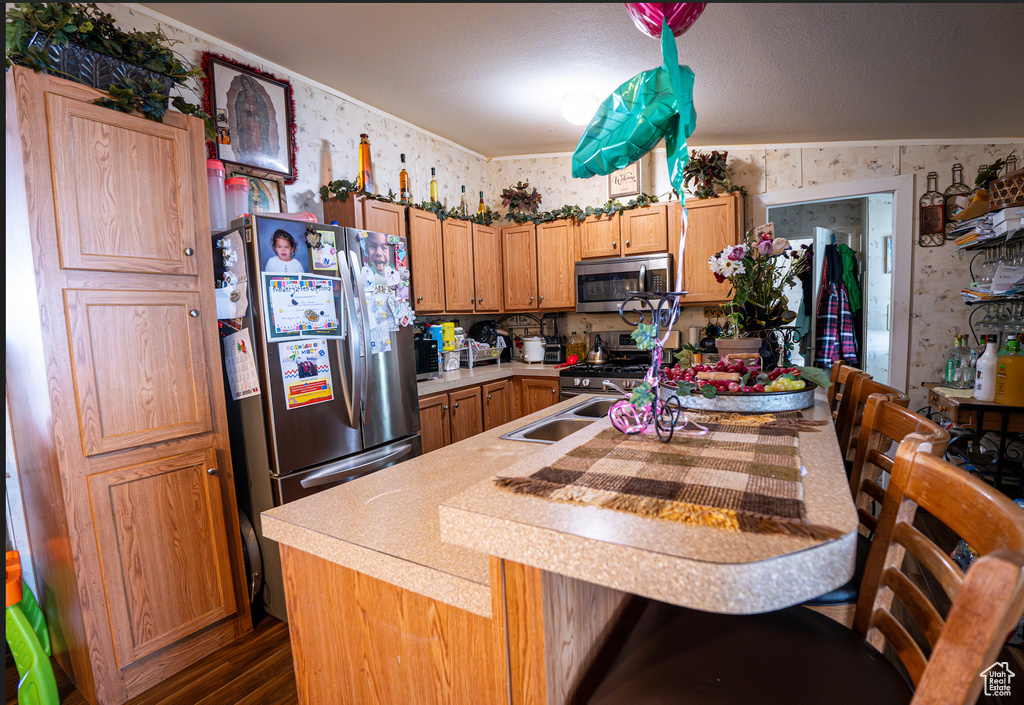 Kitchen featuring wallpapered walls, appliances with stainless steel finishes, dark wood-style flooring, light countertops, and a center island with sink