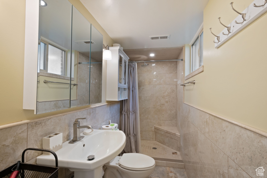 Bathroom featuring tile walls, toilet, a tile shower, wainscoting, and a sink