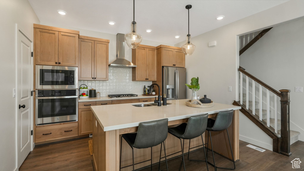 Kitchen with appliances with stainless steel finishes, wall chimney range hood, dark wood-style flooring, a kitchen island with sink, and decorative backsplash